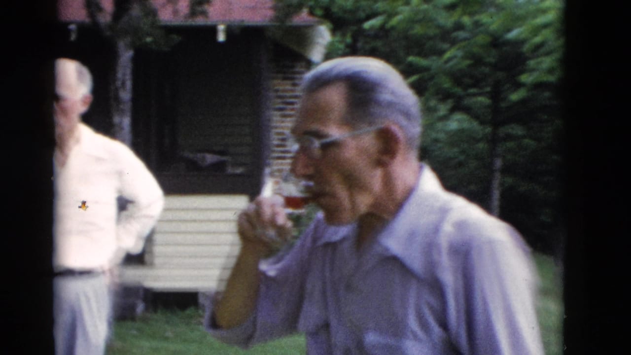 Older man drinks from a glass in the foreground as another man stands by a rustic cabin in Somerset, Kentucky, 1975.