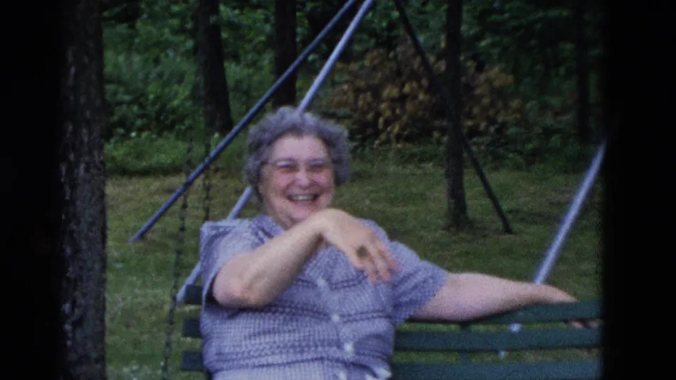 Older woman laughs while seated on a green swing in a wooded yard in Somerset, Kentucky, 1975.