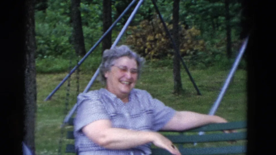 Older woman smiles from a green bench swing, with trees and lawn behind her in Somerset, Kentucky, 1975.