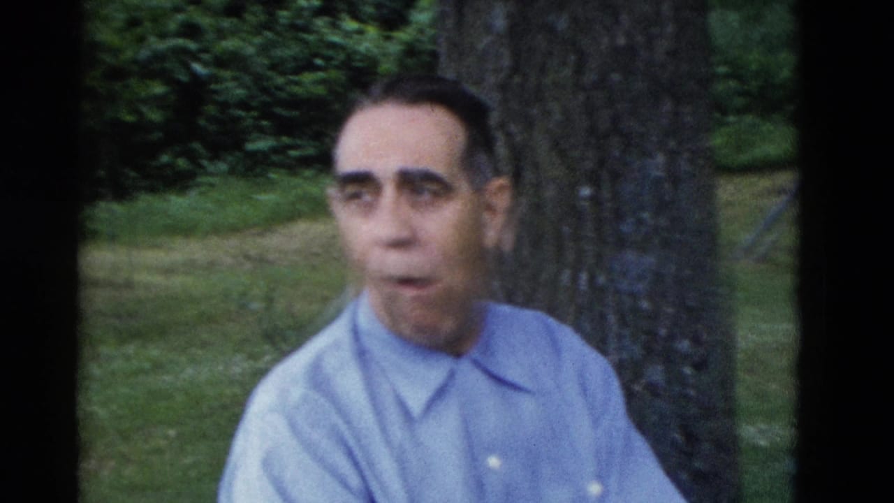 Older man in a light blue shirt sits near a tree in a grassy park.