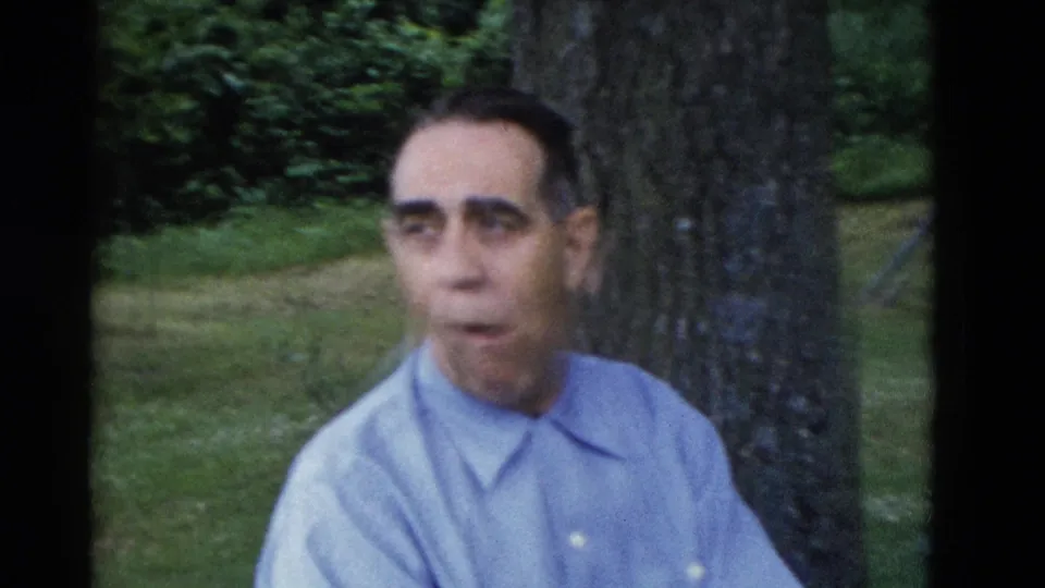 An older man in a pale blue shirt is seated beside a large tree in the wooded Somerset, Kentucky picnic setting.