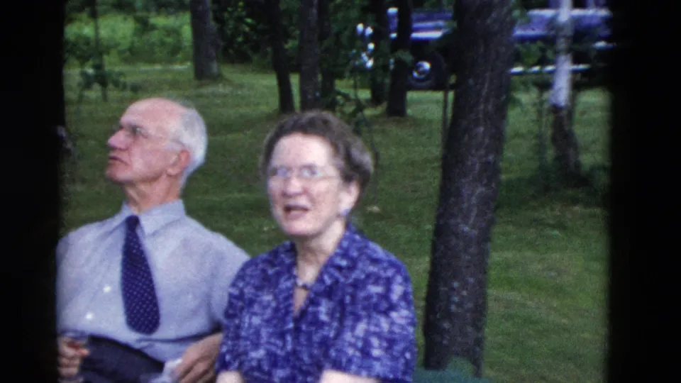 An older couple sits in the wooded picnic area, with the man in a tie holding a drink and the woman wearing glasses and a blue patterned dress.