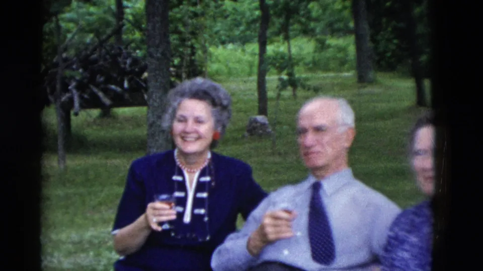 The home-movie frame shows a smiling woman in a dark jacket and an older man in a tie seated outdoors with drinks.