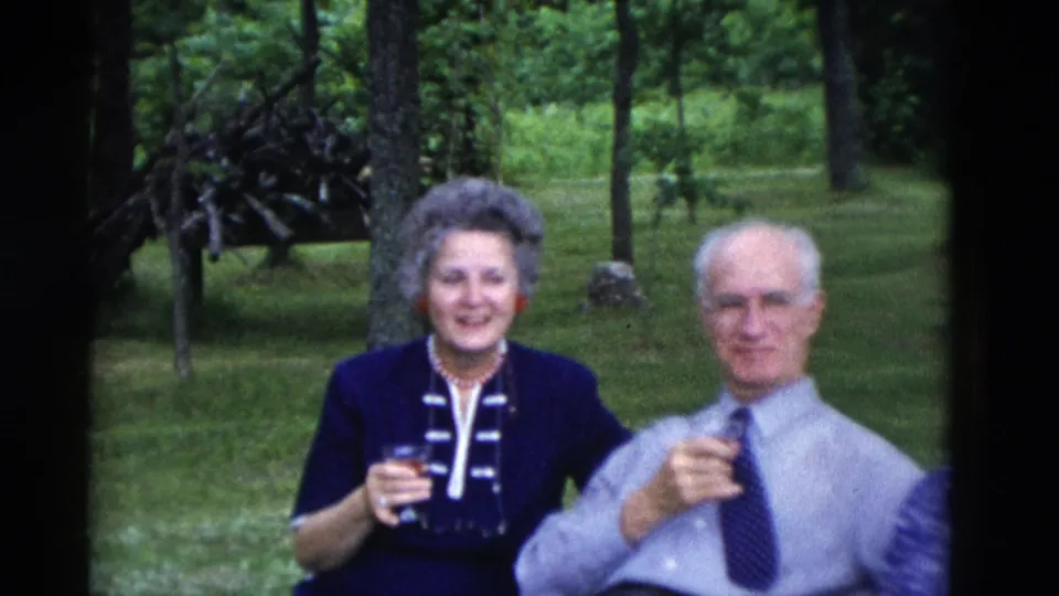In the wooded backyard scene, a woman and older man face the camera as they hold drinks during a casual gathering.