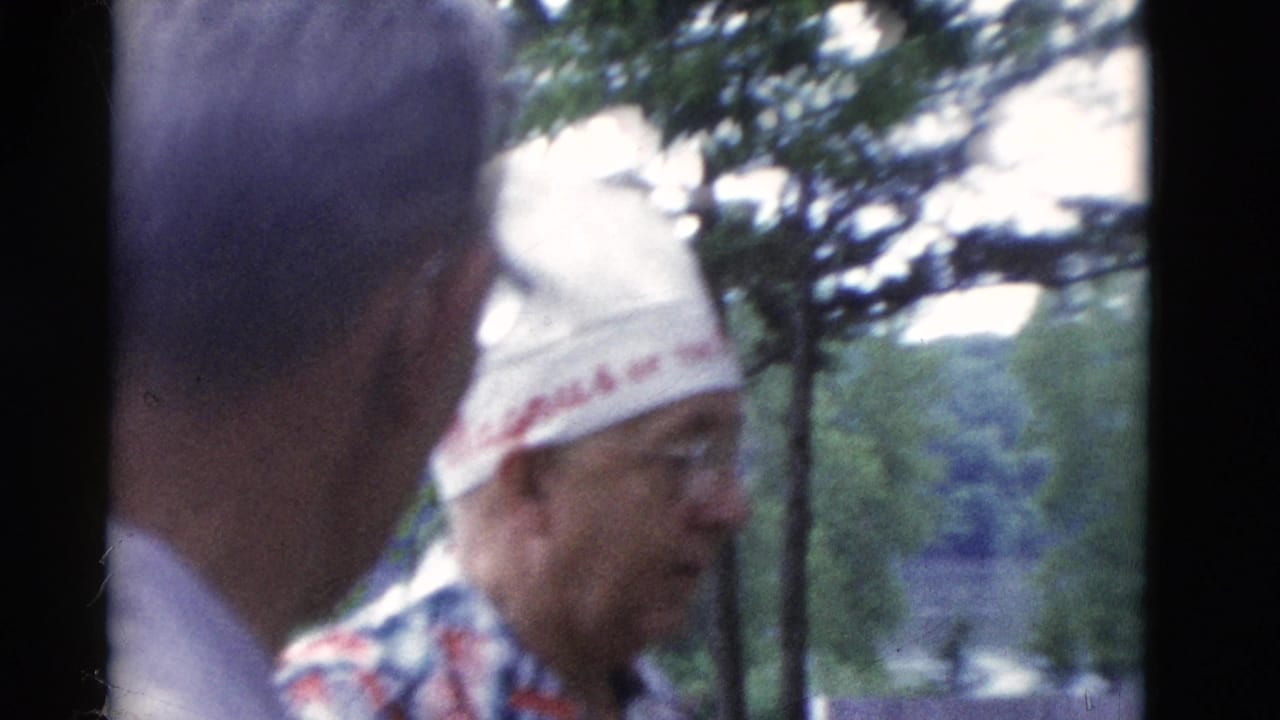 Two older men in profile outdoors, one wearing a white embroidered hat near trees.