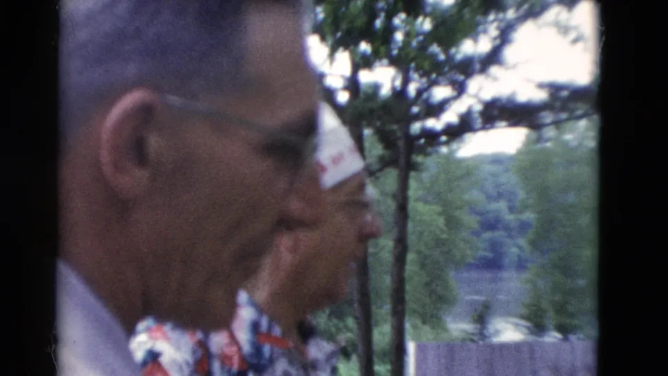 Close 8mm view of an older man with glasses beside a hat-wearing man at a wooded Somerset, Kentucky setting in 1975.