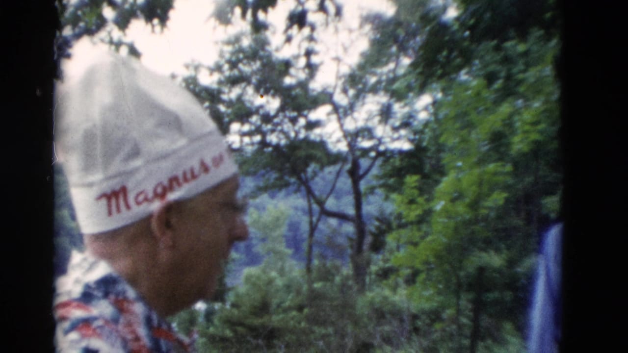 Man in a white hat and patterned shirt stands in profile outdoors near trees.
