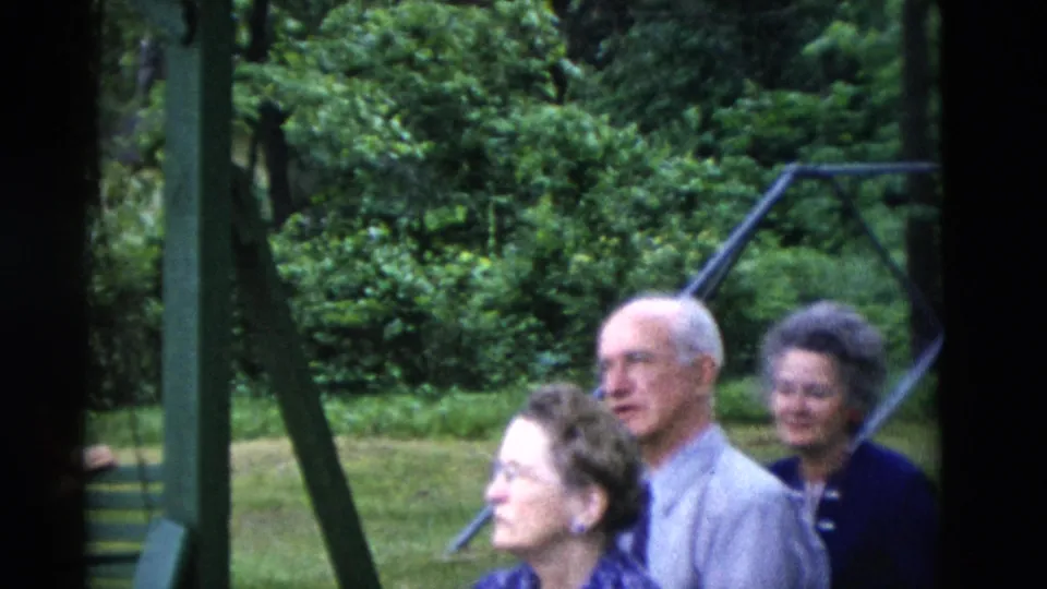 1975 Somerset, Kentucky home-movie frame showing older adults gathered outdoors beside green swing-set supports.