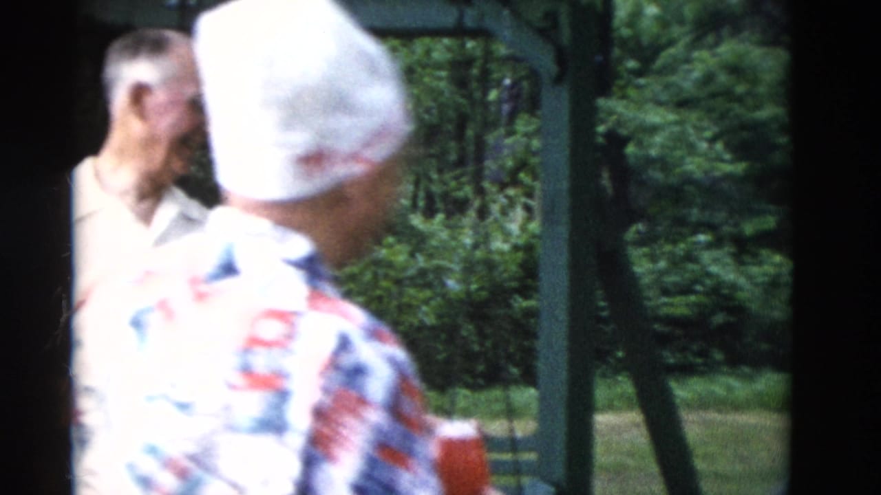 Man in white cap and patterned shirt stands near older adults in a wooded outdoor setting.