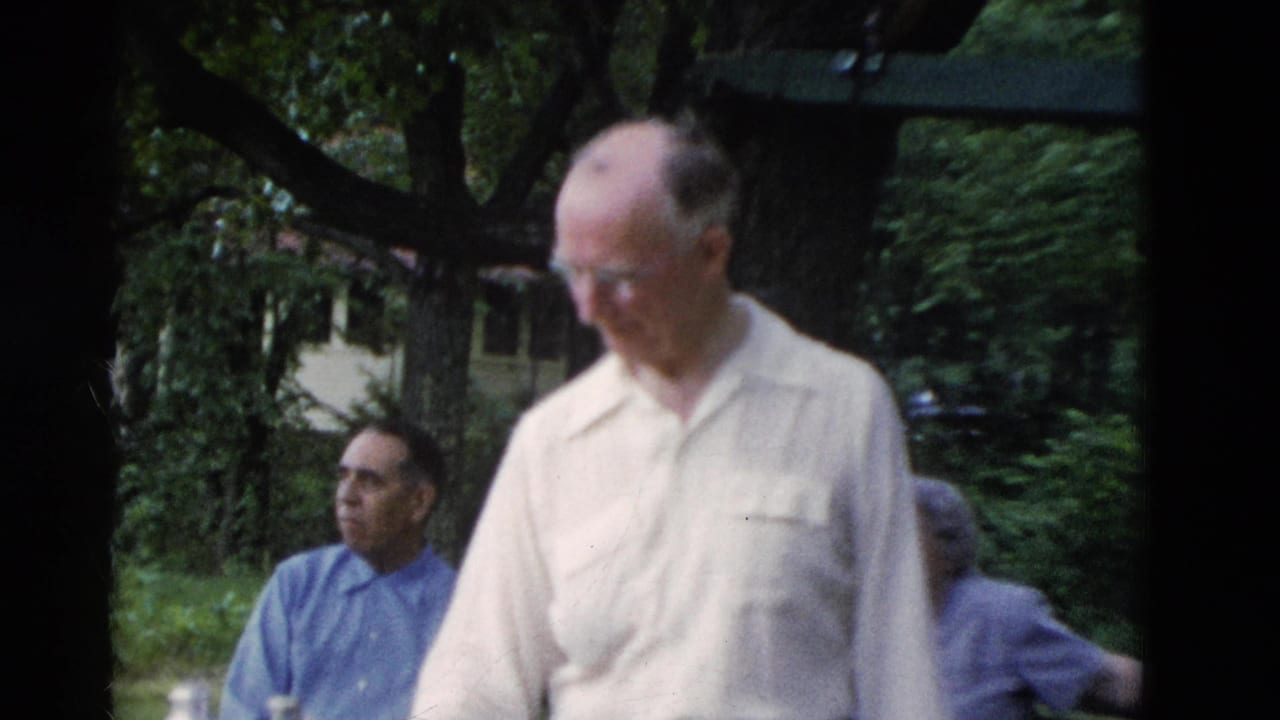 Older men outdoors near trees, with one man in a light shirt standing in the foreground.