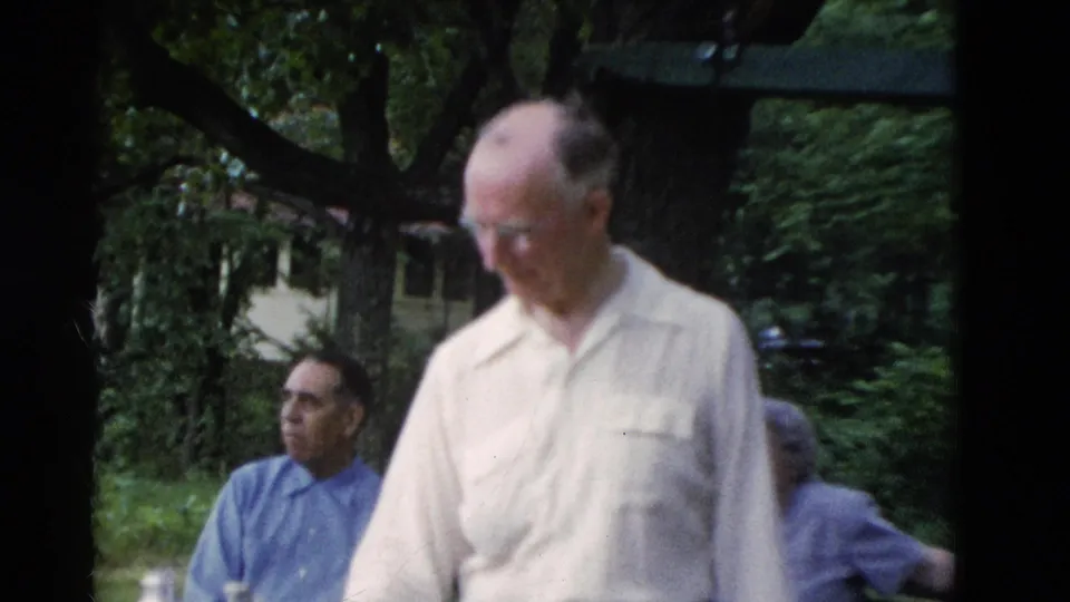 1975 Somerset, Kentucky home-movie frame showing older men gathered outdoors under trees, one in a light short-sleeve shirt near the camera.