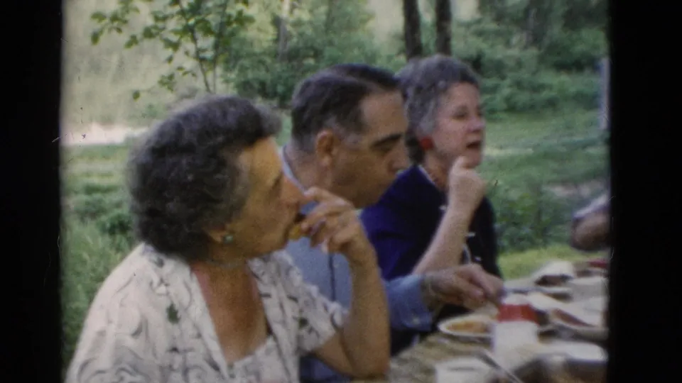 1975 Somerset, Kentucky home-movie frame showing older adults seated at a picnic table with plates, cups, and food outdoors.