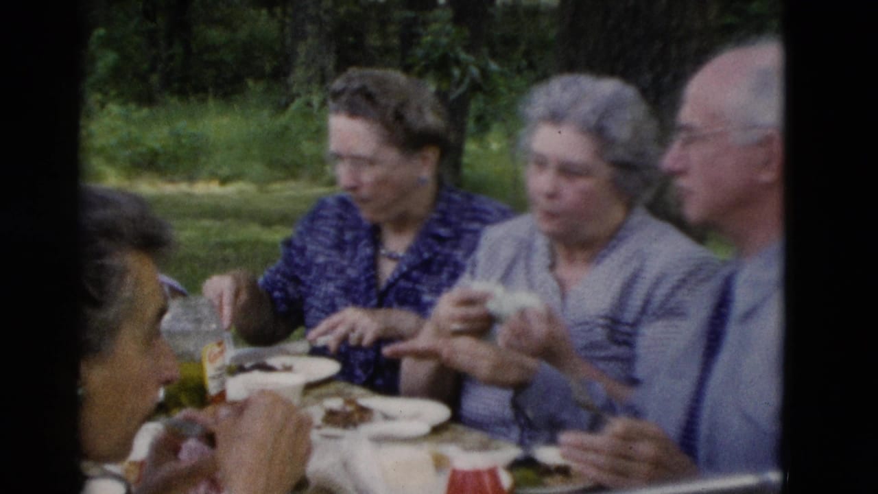 Three older adults sit at a picnic table while a woman holds food near her mouth.
