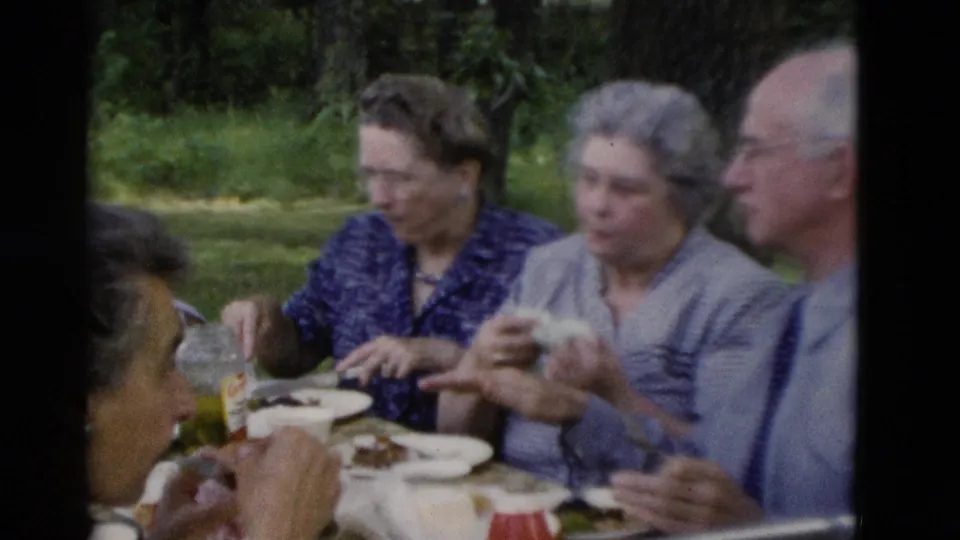 Older adults at a 1975 Somerset, Kentucky outdoor picnic, with plates, drinks and trees in the background.