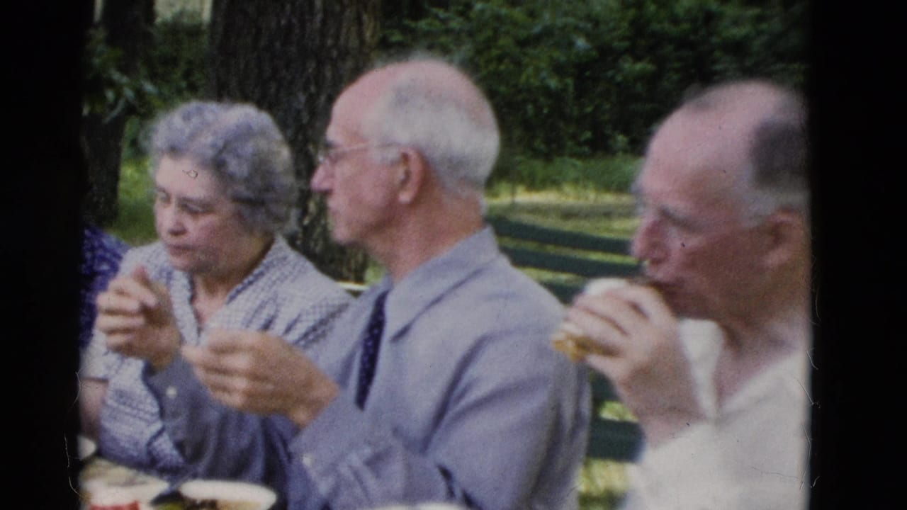 1975 Somerset, Kentucky home-movie frame showing older adults at a picnic table, with one man eating a sandwich beside a man in a tie.