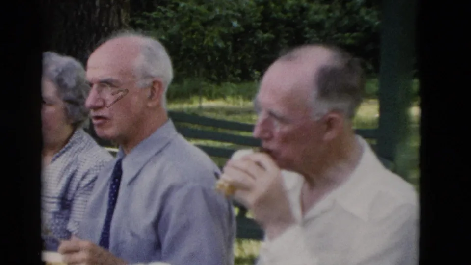 1975 8mm frame from Somerset, Kentucky showing two older men at a picnic table, one wearing a tie and another eating a sandwich.