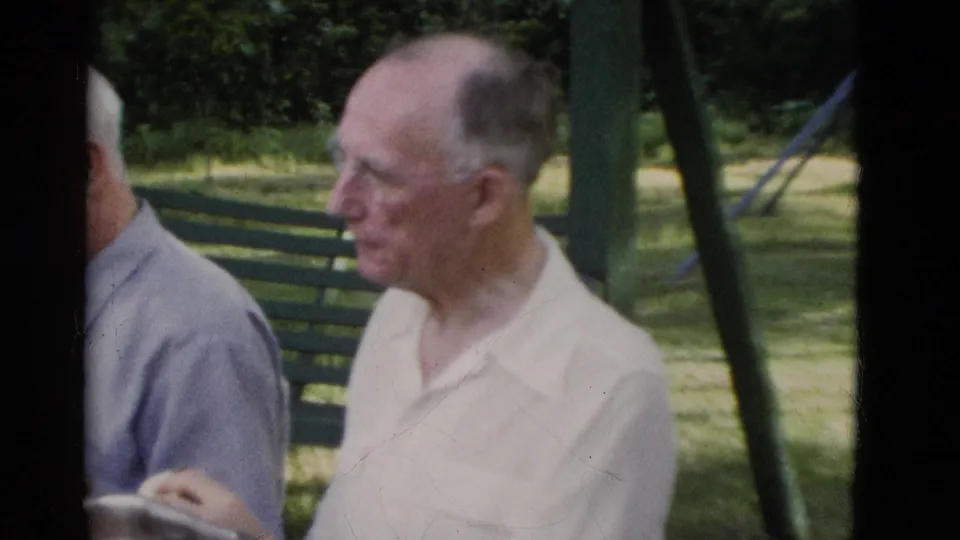 1975 Somerset, Kentucky home-movie frame showing an older man in a light short-sleeved shirt seated near a picnic table and bench.