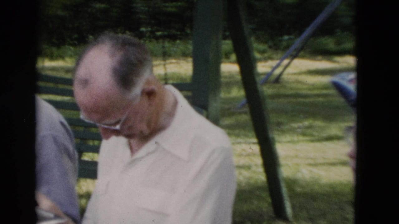 In supplied 1975 Somerset, Kentucky footage, an older man in a pale shirt and glasses sits outdoors near other adults in a grassy park area.