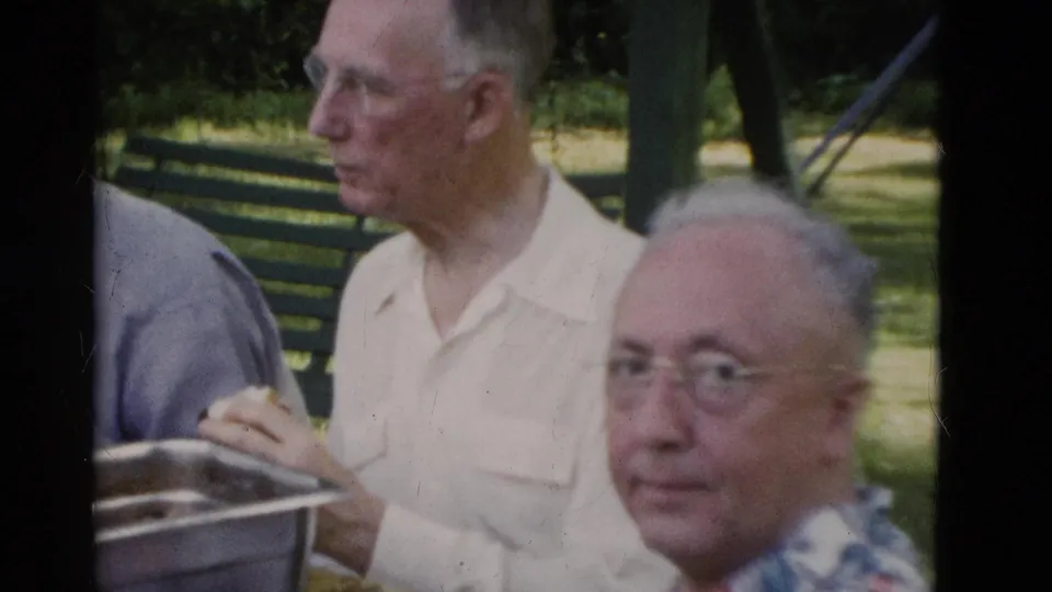 Older men sit near a metal serving pan in the supplied 1975 Somerset, Kentucky home-movie footage, with a green bench and trees behind them.