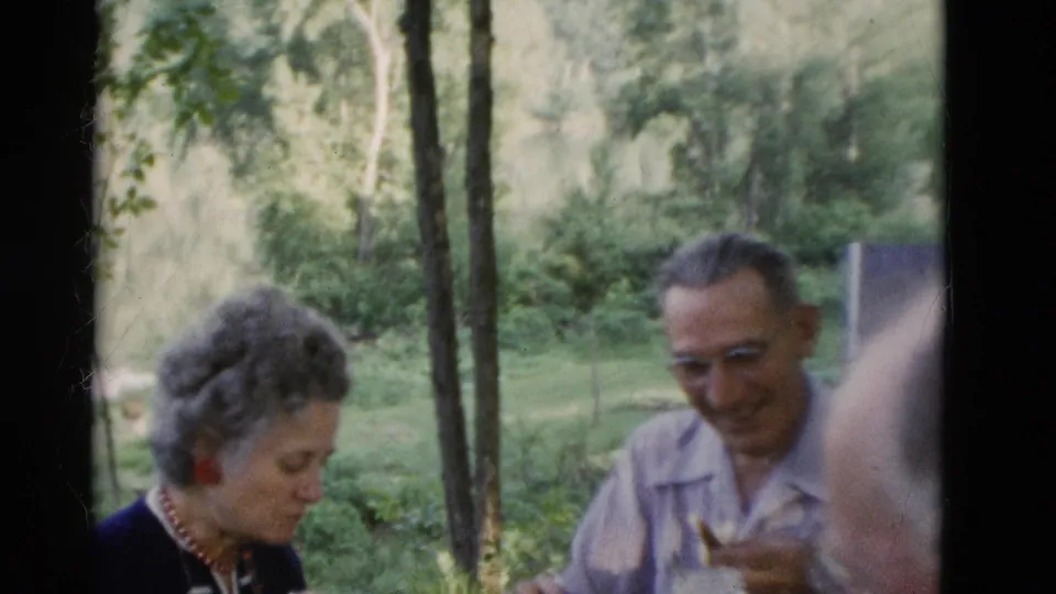 1975 Somerset, Kentucky home-movie frame showing a woman and man at an outdoor picnic table against trees and grass.