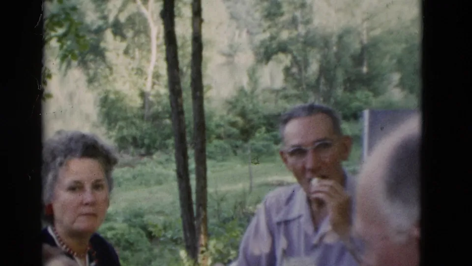 1975 Somerset, Kentucky frame showing a woman looking toward the camera and a man eating during an outdoor picnic.