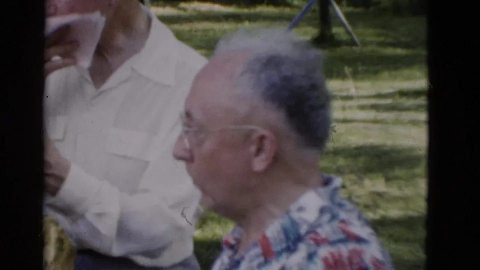 1975 Somerset, Kentucky 8mm frame showing an older man in glasses in profile with a standing adult and cup partly visible nearby.