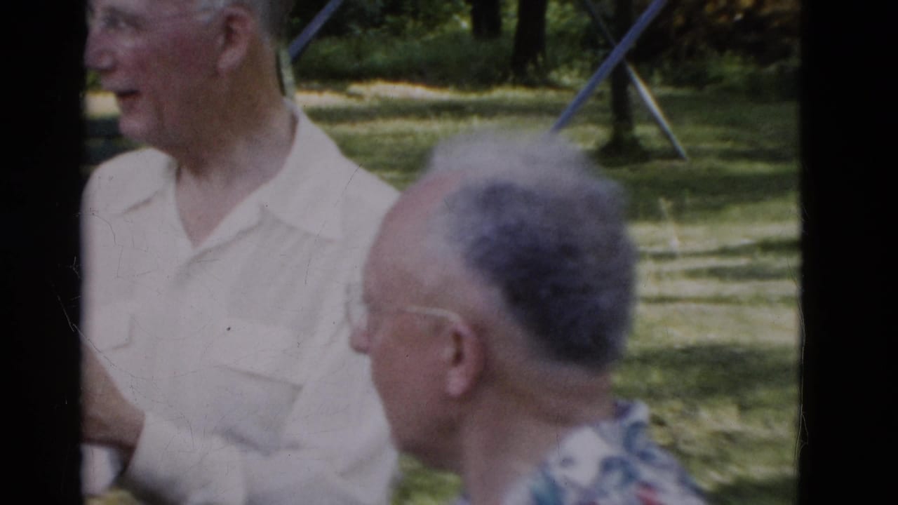 In 1975 Somerset, Kentucky footage, older men in collared shirts stand and sit near a park bench with trees and a swing set in the background.
