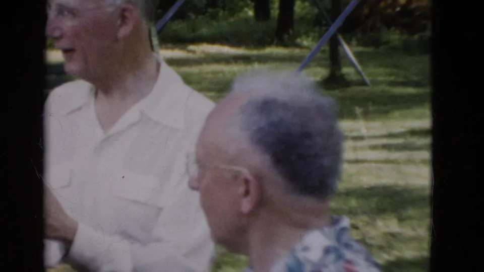 In 1975 Somerset, Kentucky footage, older men in collared shirts stand and sit near a park bench with trees and a swing set in the background.