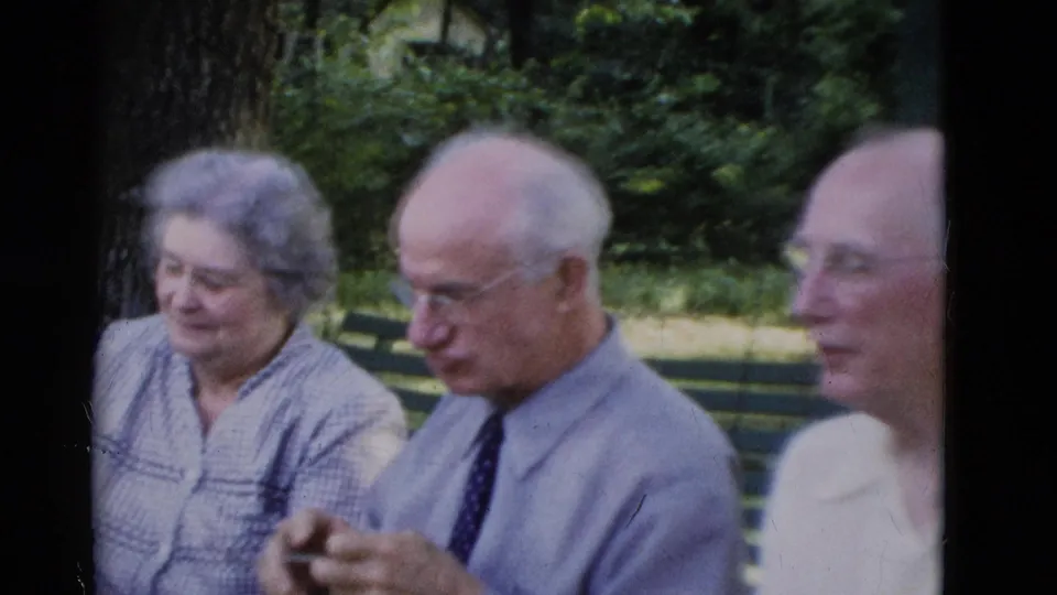 Older adults sit on a green park bench in Somerset, Kentucky, in 1975 home-movie footage, with trees and lawn behind them.