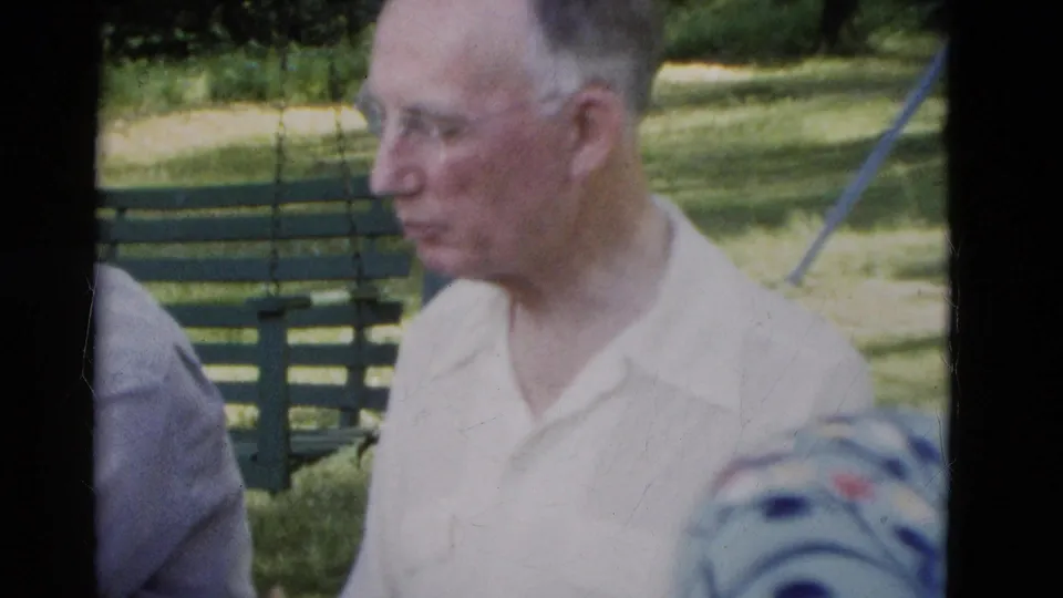 An older man in glasses and a light collared shirt stands or sits beside a green park bench in 1975 Somerset, Kentucky park footage.