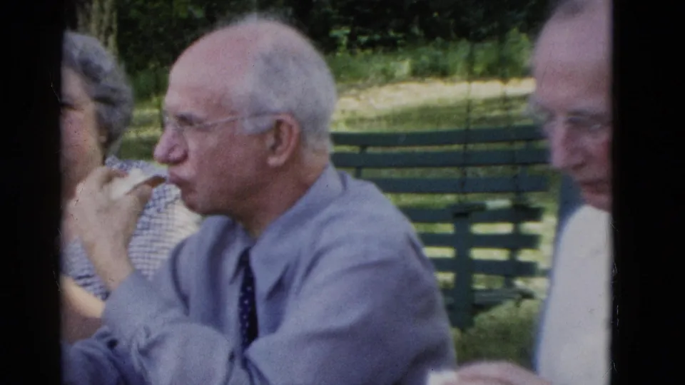 1975 Somerset, Kentucky home-movie frame of an elderly man eating a sandwich at a park bench with companions nearby.