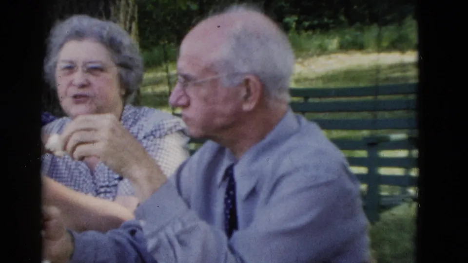 1975 Somerset, Kentucky frame of a smiling woman in a checkered dress beside an elderly man in a blue shirt during an outdoor lunch.