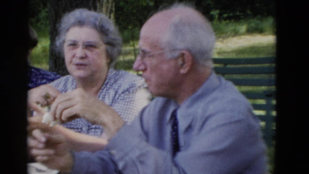 1975 Somerset, Kentucky home-movie frame of older adults at a park picnic table, with a man in a tie and a woman in a checkered top eating.