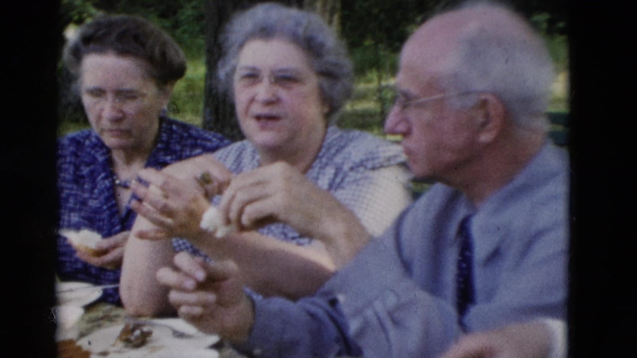 Three older adults sit at a picnic table outdoors, eating and talking.