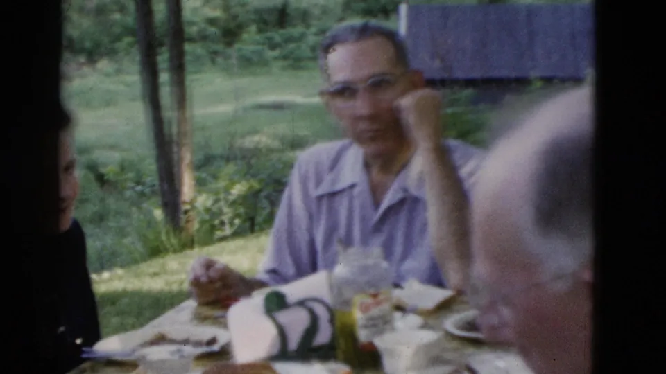 A soft handheld frame shows a man at the picnic table with plates, food, and a labeled jar in the wooded Somerset gathering.