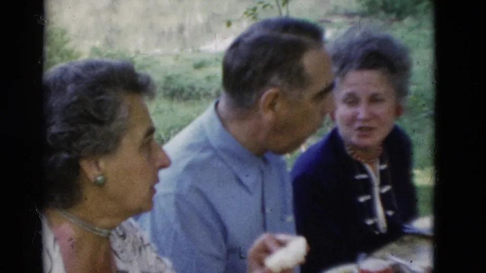 1975 Somerset, Kentucky 8mm frame of three older adults talking at an outdoor meal table in a wooded grassy area.