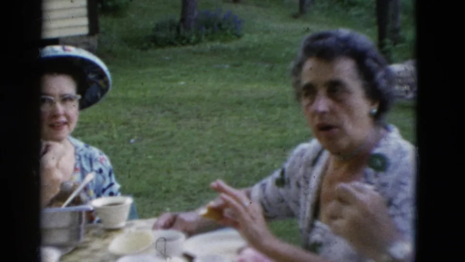 A woman in a wide-brimmed hat and glasses sits near another woman in a patterned blouse during a 1975 outdoor family meal in Somerset, Kentucky.