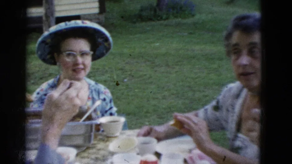 At the end of the brief 1975 Somerset, Kentucky picnic scene, a woman in a brimmed hat and glasses smiles while another woman sits beside the table.