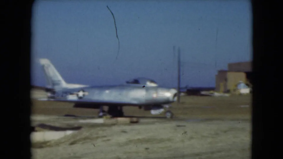 1975 Middletown, Pennsylvania 8mm frame of a silver military jet parked on an airfield with buildings in the background.