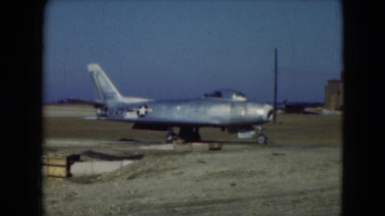Silver military jet parked on a gravel airfield under a blue sky.