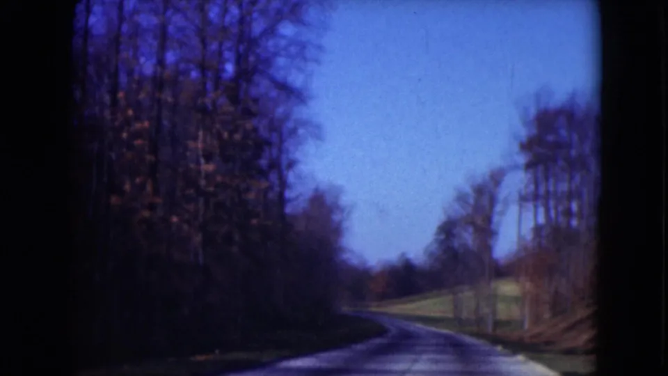 1975 Middletown, Pennsylvania 8mm frame of a wooded roadside and curving paved road seen from a handheld home-movie viewpoint.