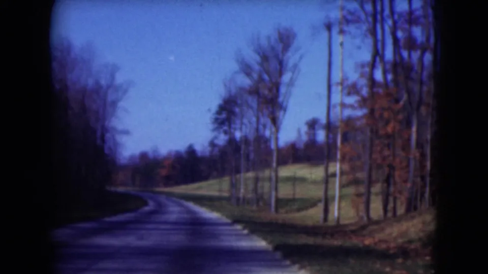 1975 8mm frame from Middletown showing a paved rural road receding toward a bend through leafless roadside trees.