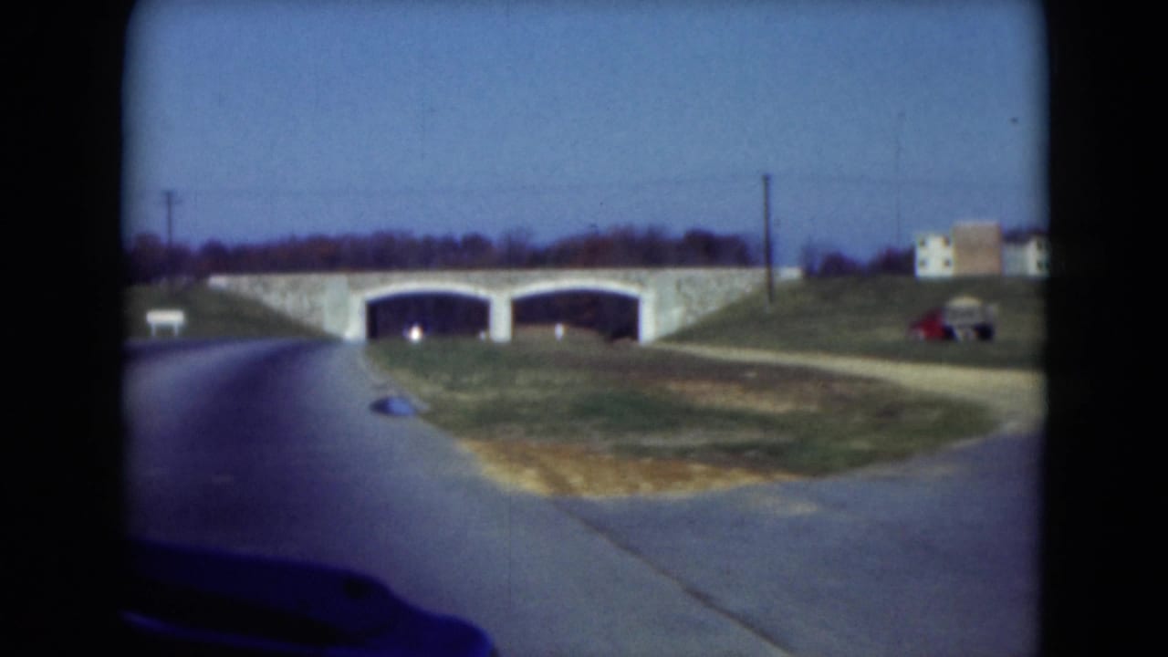 A moving-car view in Middletown, Pennsylvania shows the road curving toward a two-arch bridge with a red vehicle on the right shoulder.