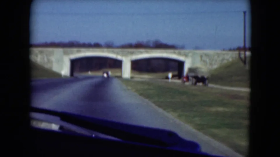 Closer 8mm view of the roadway leading to the double-arch stone bridge, with vehicles or equipment visible near the right shoulder.