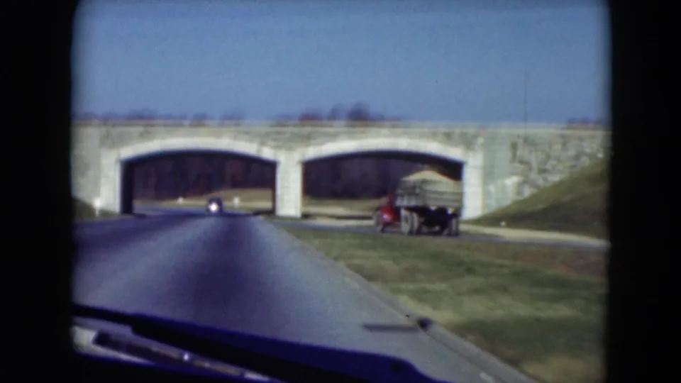 Near the bridge, the moving-car view shows a parked truck or trailer on the right shoulder beside the stone arches and grassy slope.