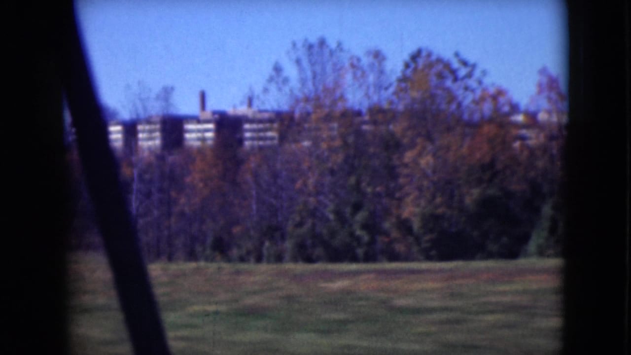 Grassy field and autumn trees with distant brick buildings seen through a vehicle window.