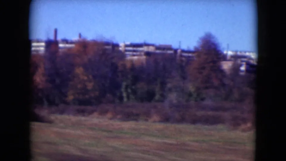 Middletown, Pennsylvania home-movie frame showing grass, fall trees, and distant institutional-looking brick buildings through a vehicle window.