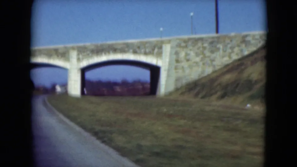 1975 Middletown, Pennsylvania home-movie frame of a stone arch bridge over a road, with grassy embankments and clear sky.