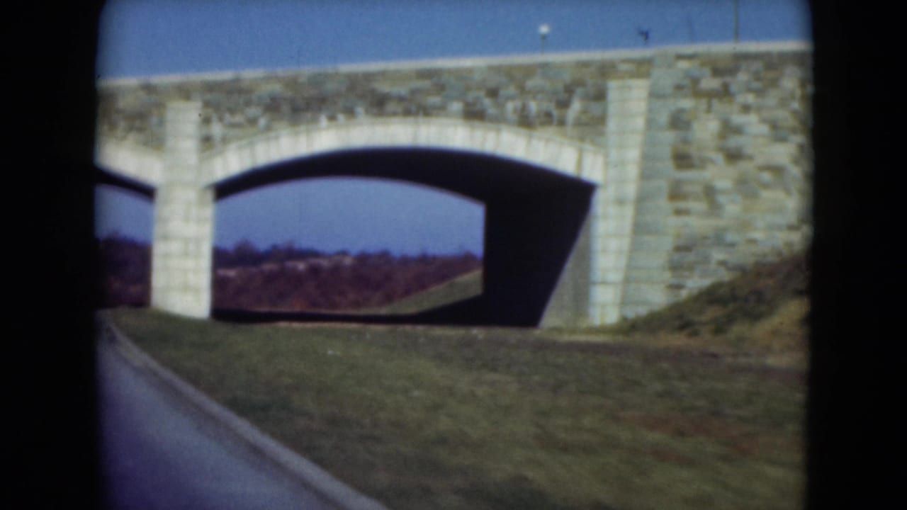 The moving 8mm view nears the masonry bridge arch, with the road edge, grass, blue sky, and tree line visible.