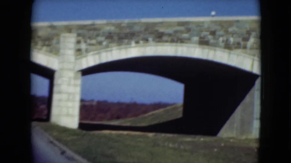 A close 1975 home-movie view of the bridge arch shows pale masonry, dark underpass shadow, grass, and open sky.
